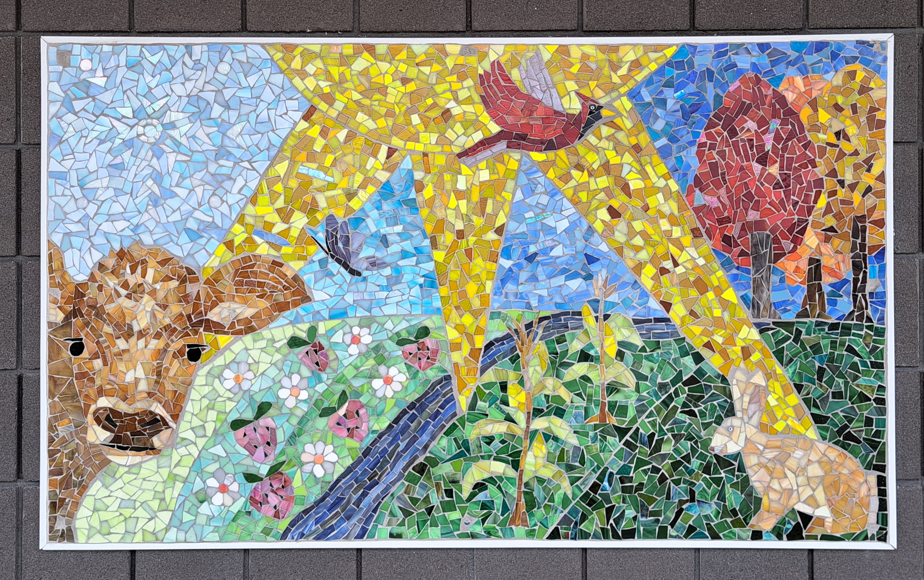 Large sun with rays dividing the mural into 4 season sections. Spring has the cute face of a brown baby calf and light blue sky with iridescent white snowflake. Summer shows pink strawberries growing in a field with a brighter blue sky and purple butterfly. Summer shows corn stalks growing in a medium green field with blue sky and red cardinal flying. Fall shows three colorful trees in a dark blue sky, and a tan rabbit in the dark green grass.