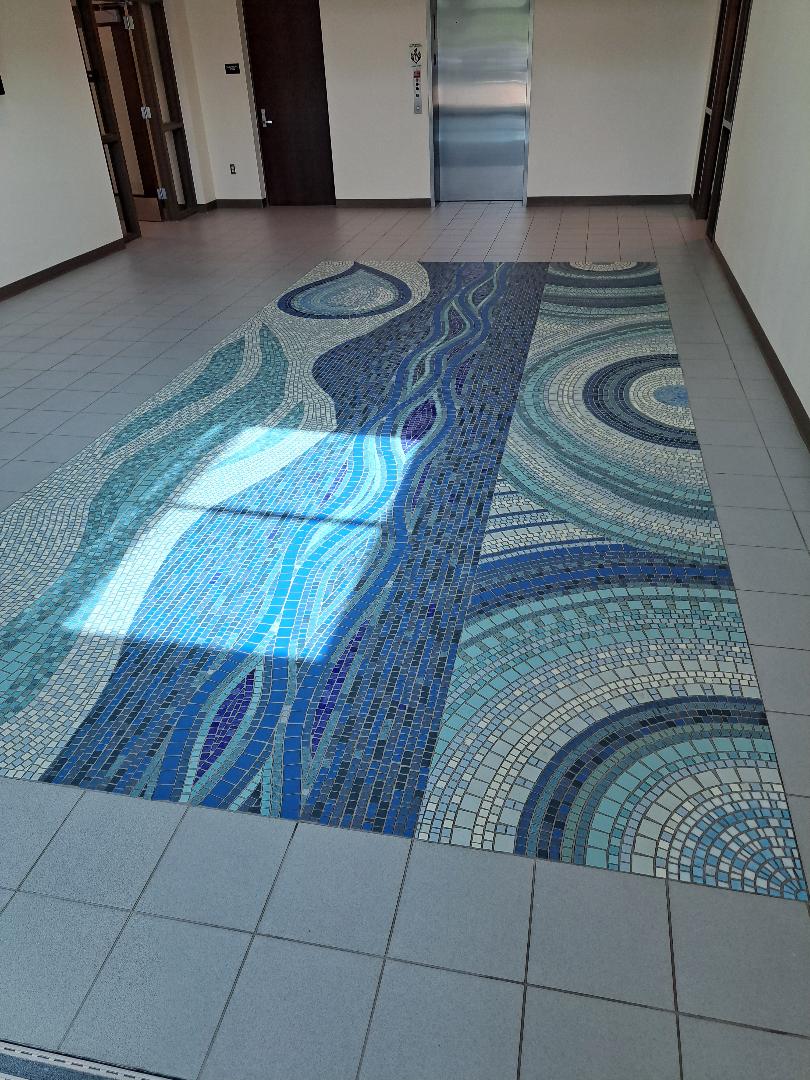 Blue mosaic tiles showing a large water droplet, flowing water in a stream, circular water ripples, in a tile floor inset.